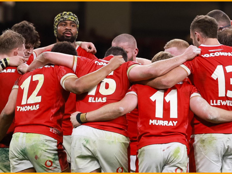 Wales players in a huddle in muddy red home kit, with Thomas, Elias, Murray and Llewellyn on their backs