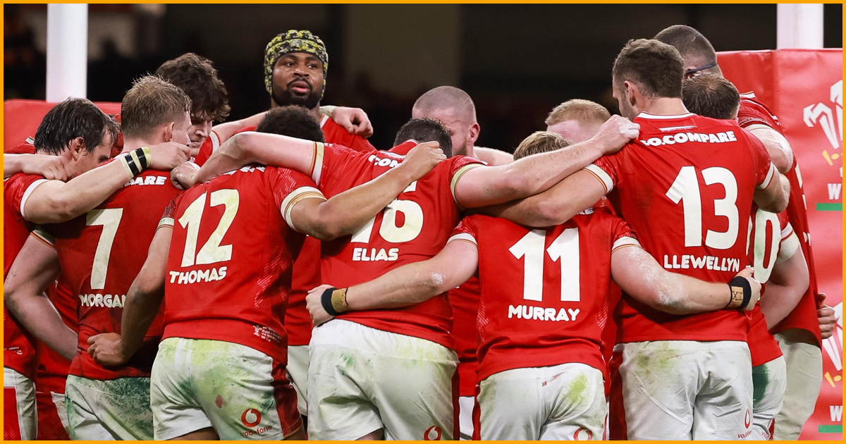 Wales players in a huddle in muddy red home kit, with Thomas, Elias, Murray and Llewellyn on their backs